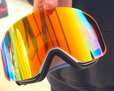 A close-up shot of a person holding a pair of custom Skoggl ski and snowboard goggles created for Corona Cero. The goggles feature a large, rimless-style cylindrical lens with a vibrant orange and yellow mirror finish that reflects a rainbow of blue and green at the edges. The frame is a crisp matte white.