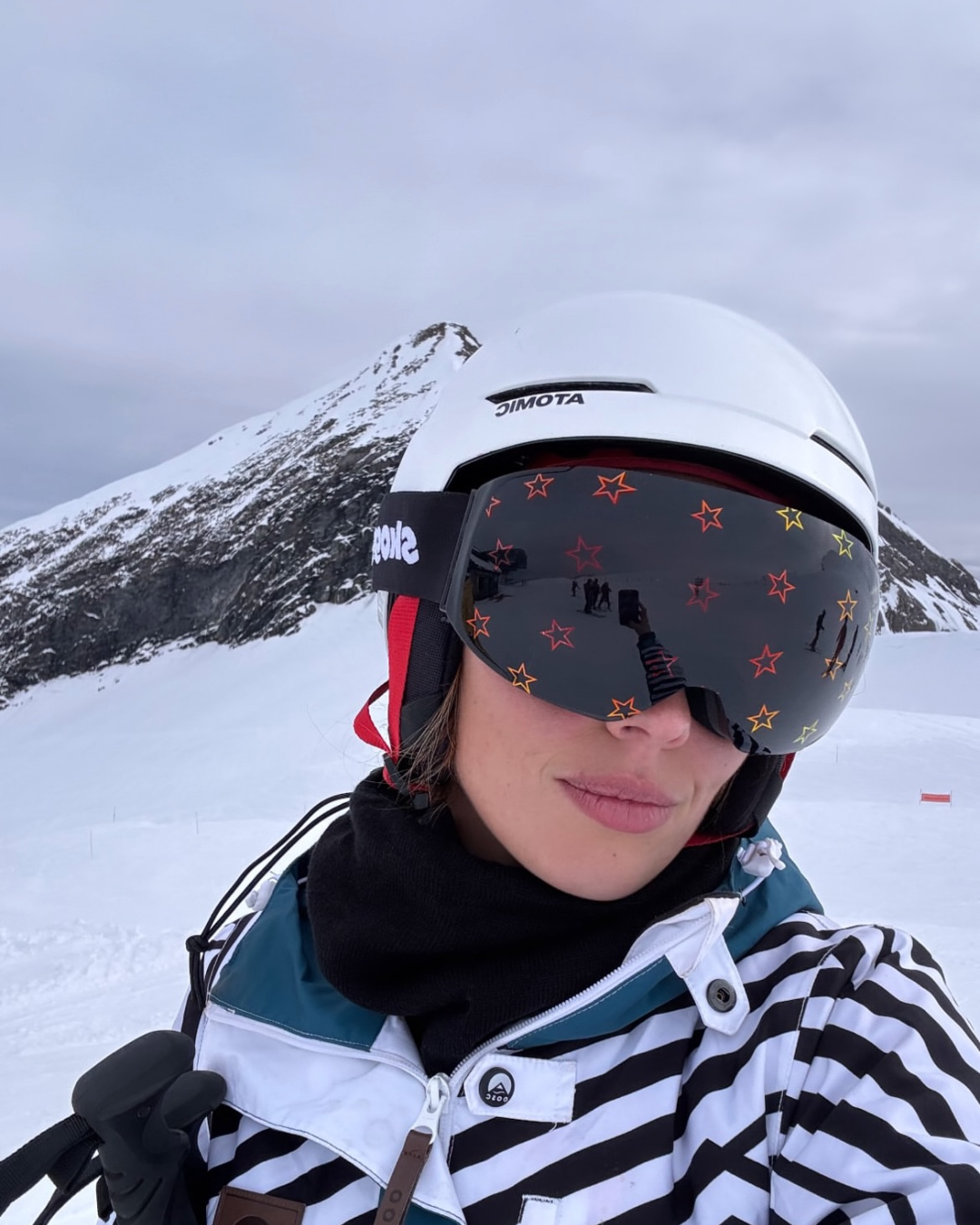 A close-up selfie of DJ Lulah Francs on a snowy mountain slope. She is wearing a white Atomic ski helmet and Skoggl goggles featuring a dark lens with orange and yellow star outlines. She is dressed in a black-and-white striped ski jacket with a black neck gaiter, with a snow-covered mountain peak visible in the background under an overcast sky.