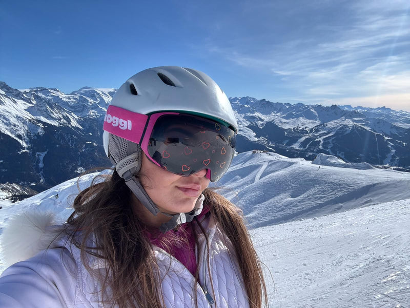 A woman wearing pink Skoggl ski goggles with heart-shaped designs on the lens stands at the top of the Swiss Alps, with a snowy mountain range stretching behind her.