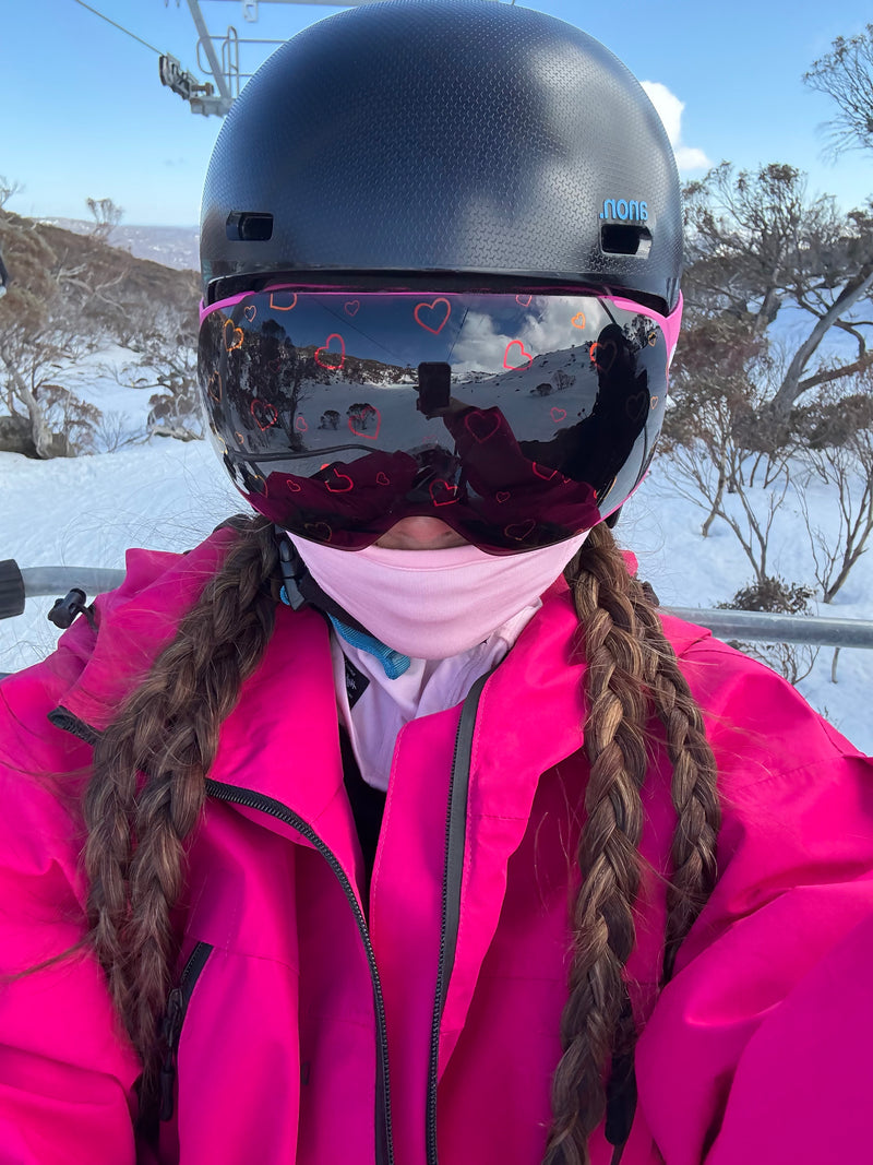 Person in pink snow gear wearing a helmet and Skoggl heart-design goggles, sitting on a chairlift with snowy mountains and a bright blue sky in the background