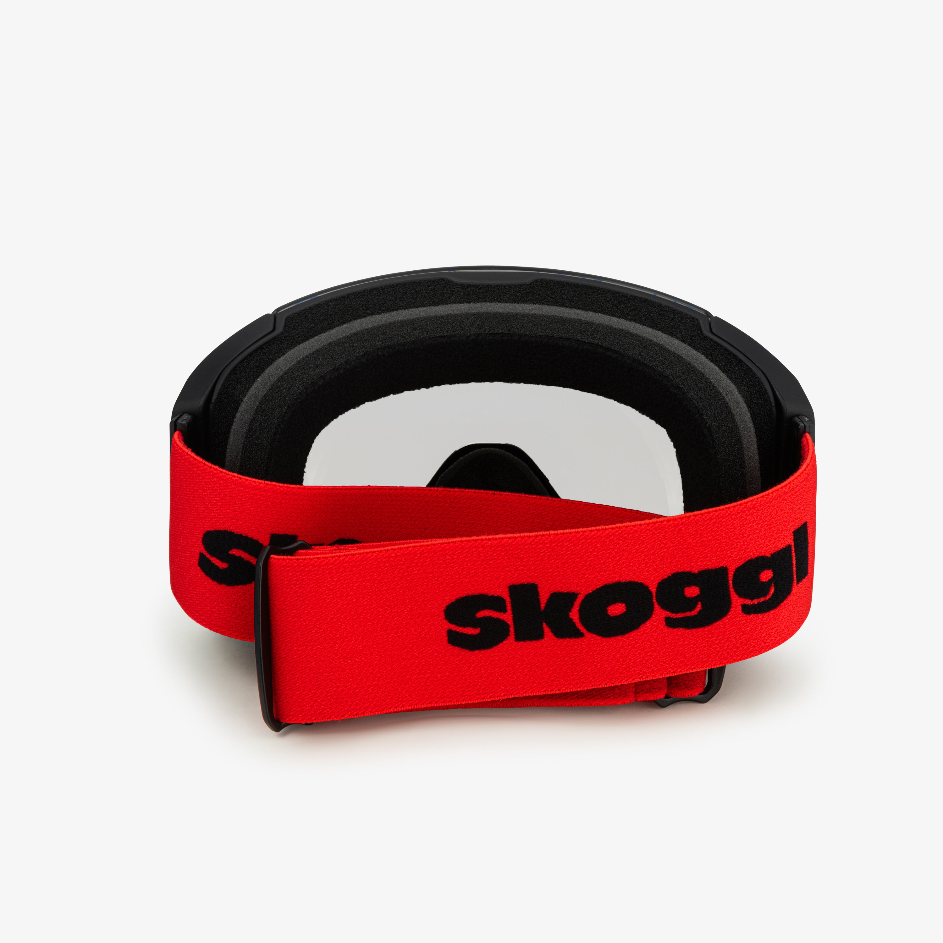 Black ski goggles with a red wide strap featuring the word 'Skoggl,' shown from the back. The clear lenses are visible through the frame, with black cushioning around the eyes.