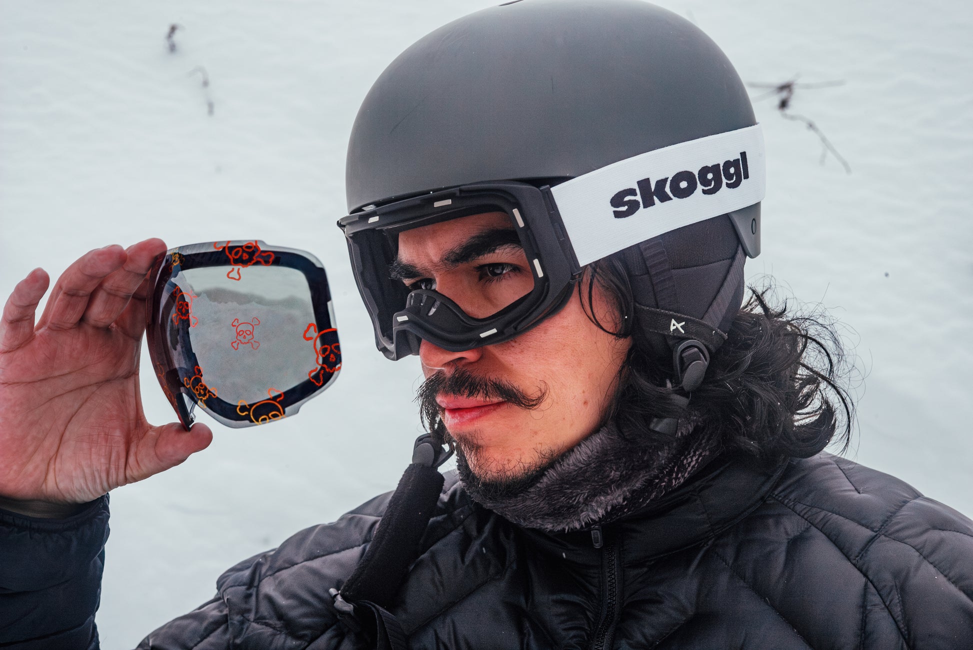 Man wearing black and red Skoggl ski goggles with interchangeable magnetic skull design pattern lens on snowy mountain