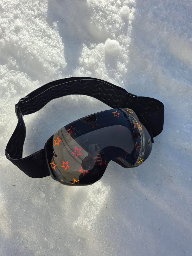 Skoggl custom children's snowboard and ski goggles with reflective star-print lenses and non-slip strap, displayed on snow.