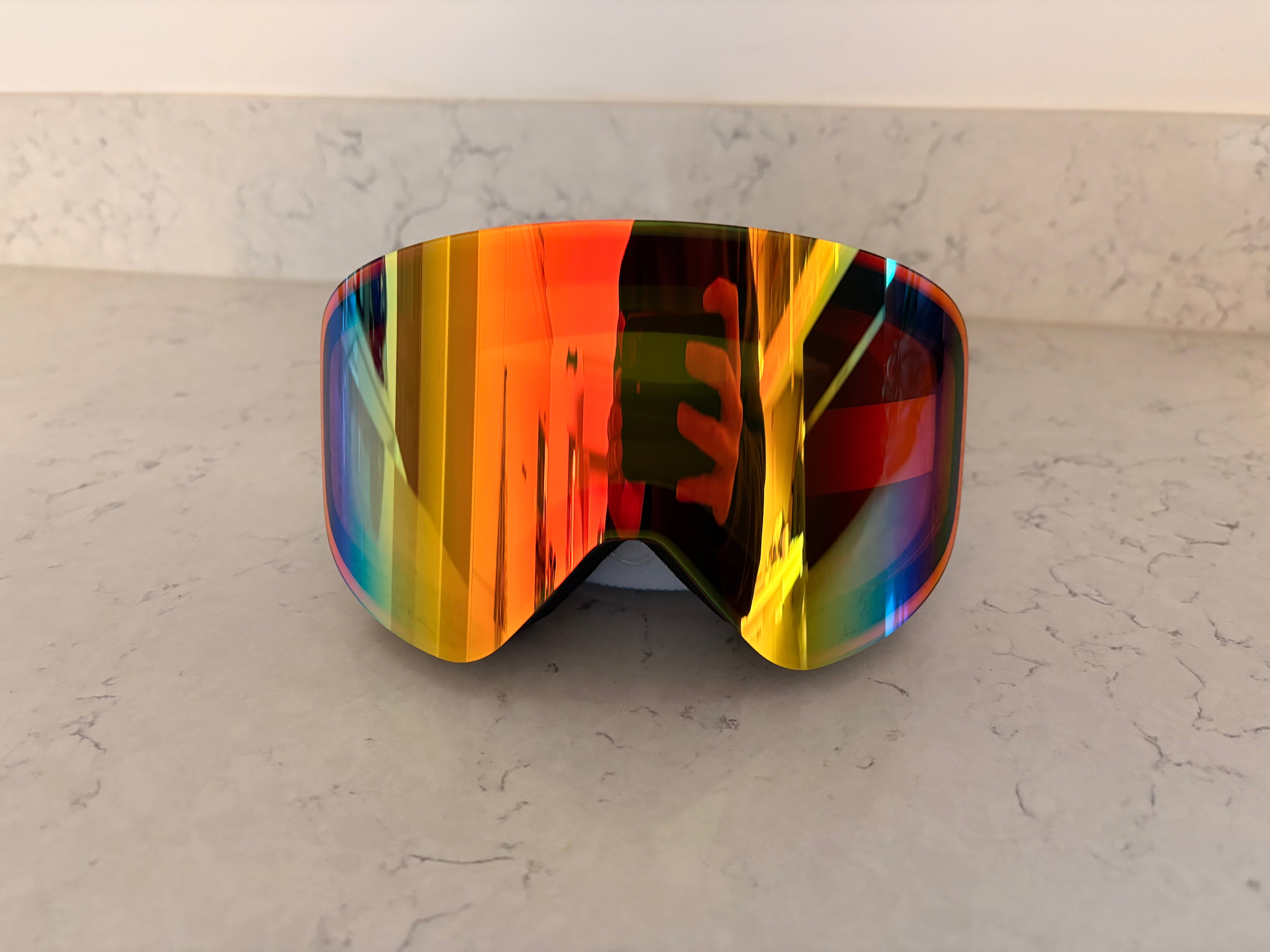 A direct front-facing shot of custom ski goggles created through a partnership between Skoggl and Corona Cero. The oversized, rimless mirrored lens dominates the frame, showcasing a striking "fire" color spectrum that transitions from bright orange and yellow in the center to deep purple and blue at the edges. The lens surface reflects a warm interior and the silhouette of the person taking the photo.