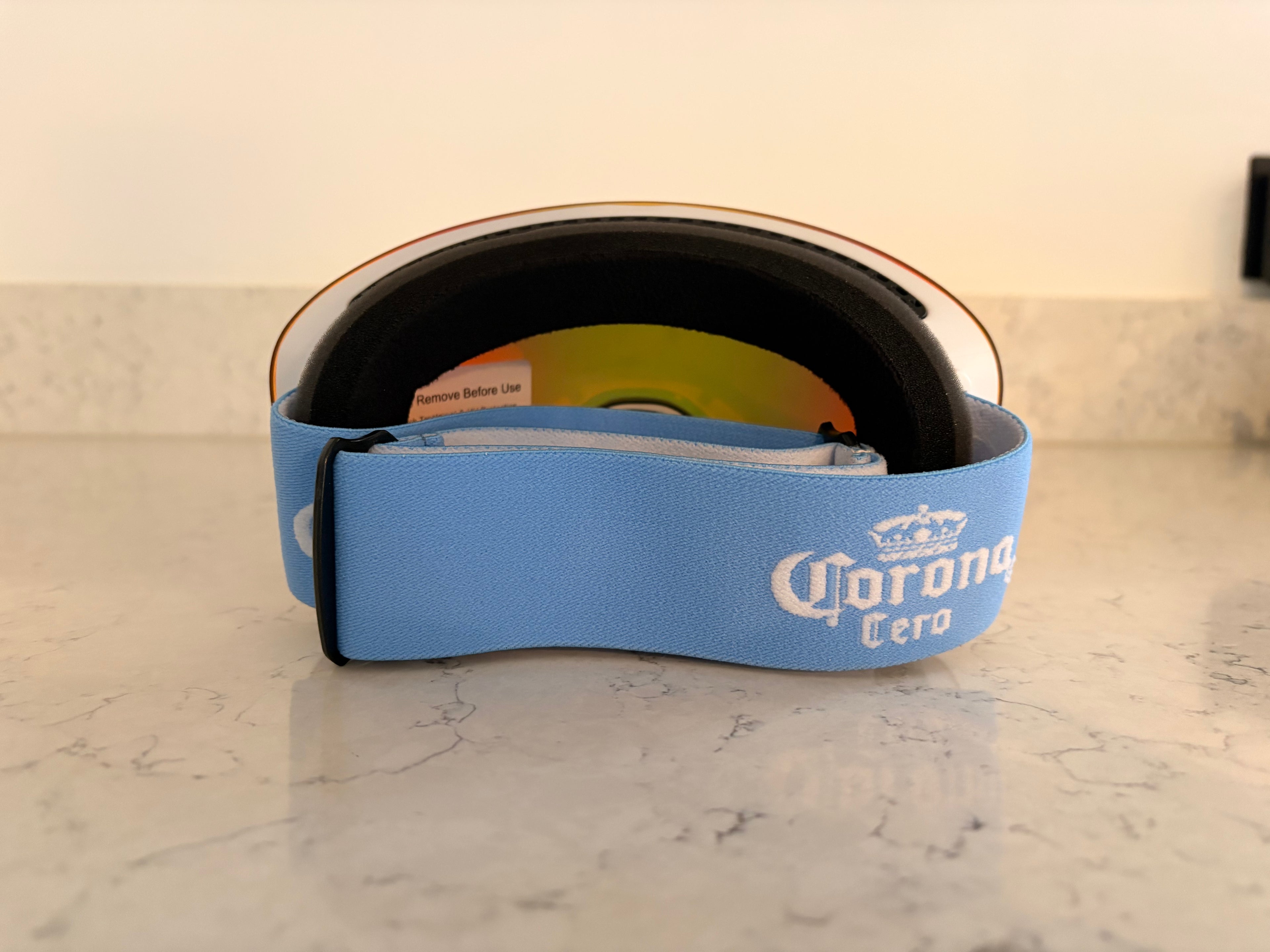 A rear view of custom ski goggles resting on a white marble surface. The thick, light blue elastic strap is prominently displayed, featuring the "Corona Cero" logo embroidered in white. The black foam padding and the interior of the orange-tinted lens are visible, including a small "Remove Before Use" sticker.
