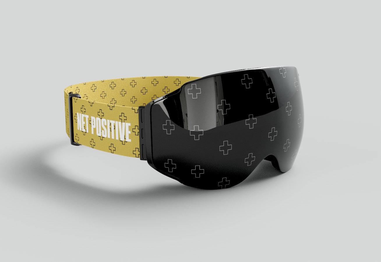 Three-quarter view of Skoggl x Net Positive custom ski and snowboard goggles, showing the black magnetic lens with an etched "plus" sign pattern and a high-visibility yellow strap featuring bold "NET POSITIVE" typography.