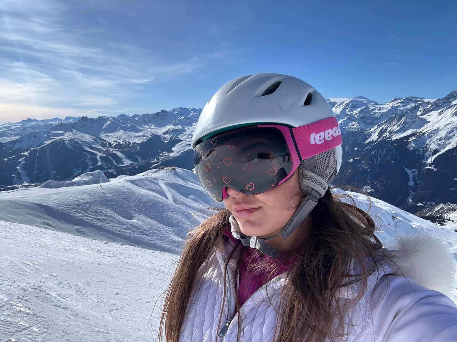 A close-up selfie of a woman on a sunny ski slope wearing a white helmet and custom Skoggl ski goggles. The goggles have a bright pink strap with a white Skoggl logo and a dark magnetic lens decorated with small, delicate pink heart outlines.
