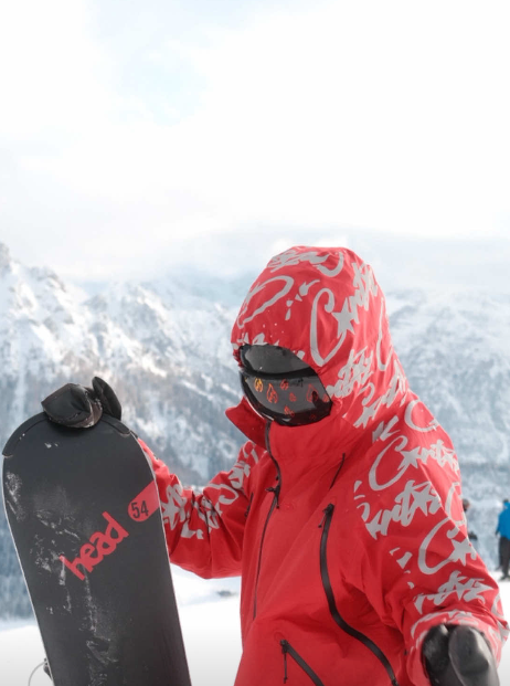 A snowboarder in a vibrant red technical jacket and hood stands against a snowy mountain backdrop, holding a black "Head" snowboard. They are wearing custom Skoggl goggles featuring a magnetic lens with a repeating orange fire flame pattern.