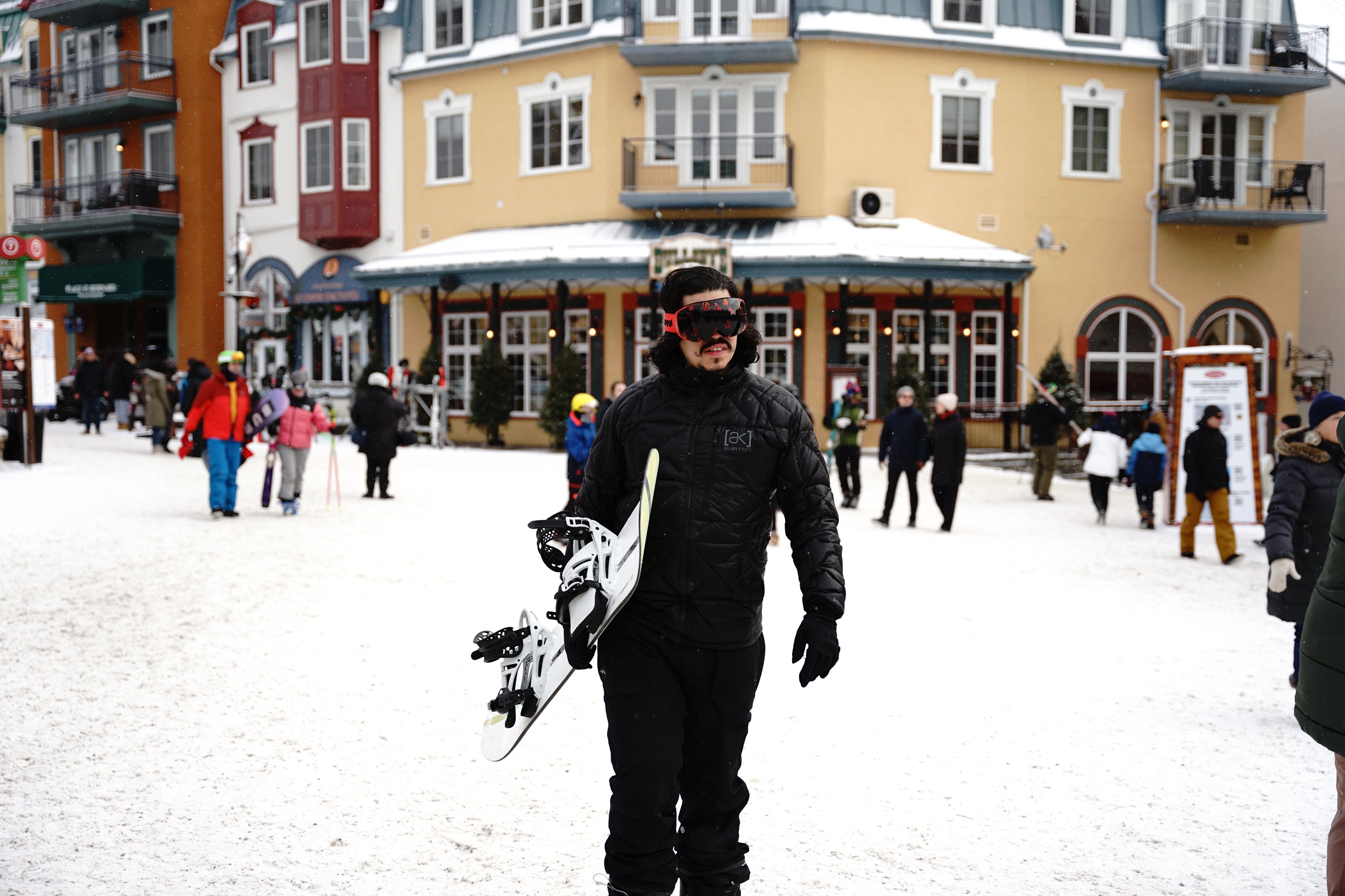 A snowboarder stands in a snowy mountain village square holding a white snowboard. He is wearing a black puffer jacket and Skoggl custom ski goggles featuring a vibrant red frame and a dark magnetic lens with a fire-patterned design. In the background are multi-story resort buildings with yellow and red facades and other visitors walking through the snow.