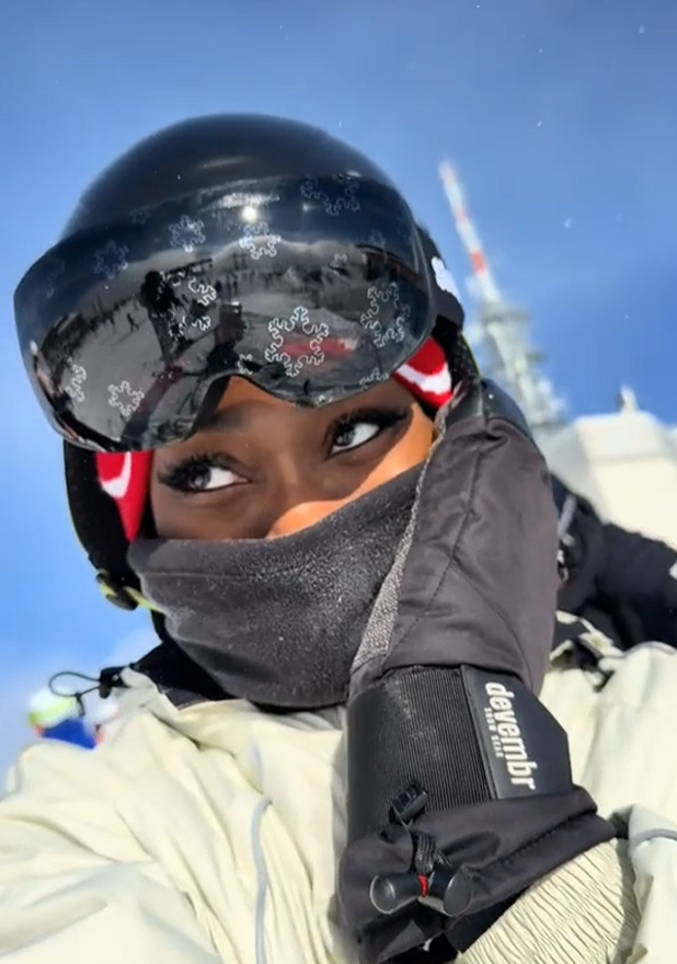 A close-up selfie of a person wearing winter gear against a clear blue sky. They are wearing a black ski helmet, a black face mask, and custom Skoggl black magnetic ski goggles featuring a white snowflake pattern across the lens. A red and white beanie is visible under the helmet, and a "devembr" branded glove is held up near the face