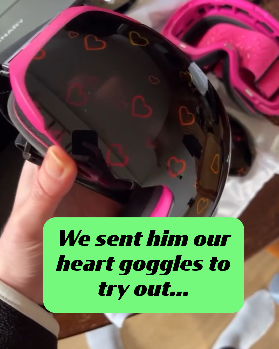 A close-up hand-held shot of a pair of Skoggl ski and snowboard goggles with a pink frame and dark lenses featuring a pattern of orange and red hearts. A green text box reads, "We sent him our heart goggles to try out...". These custom ski and snowboard goggles were sent to paralympian Matthew Hamilton