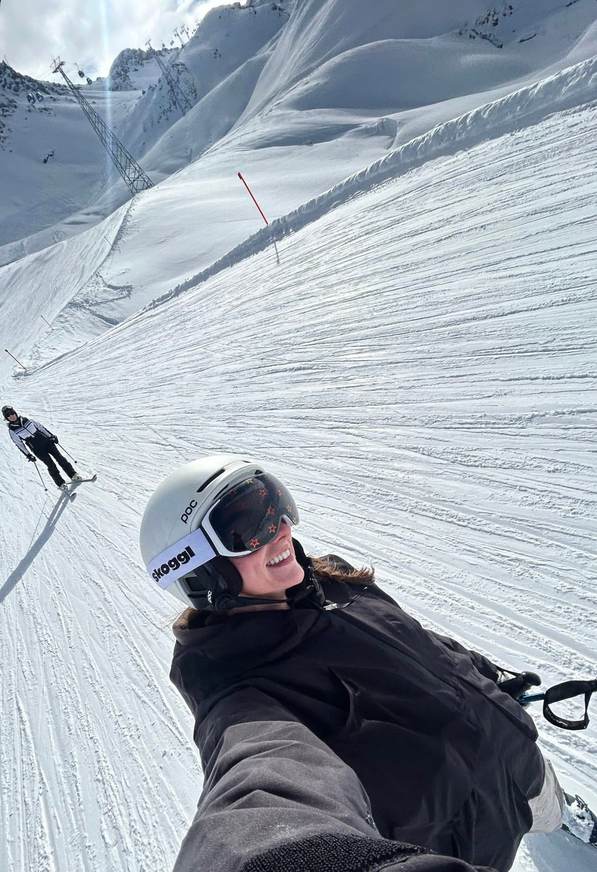 A woman smiles while skiing down a groomed mountain run, wearing a white "POC" helmet and custom Skoggl goggles. These goggles feature a white strap with a black logo and a dark lens adorned with small, repeating red star outlines.