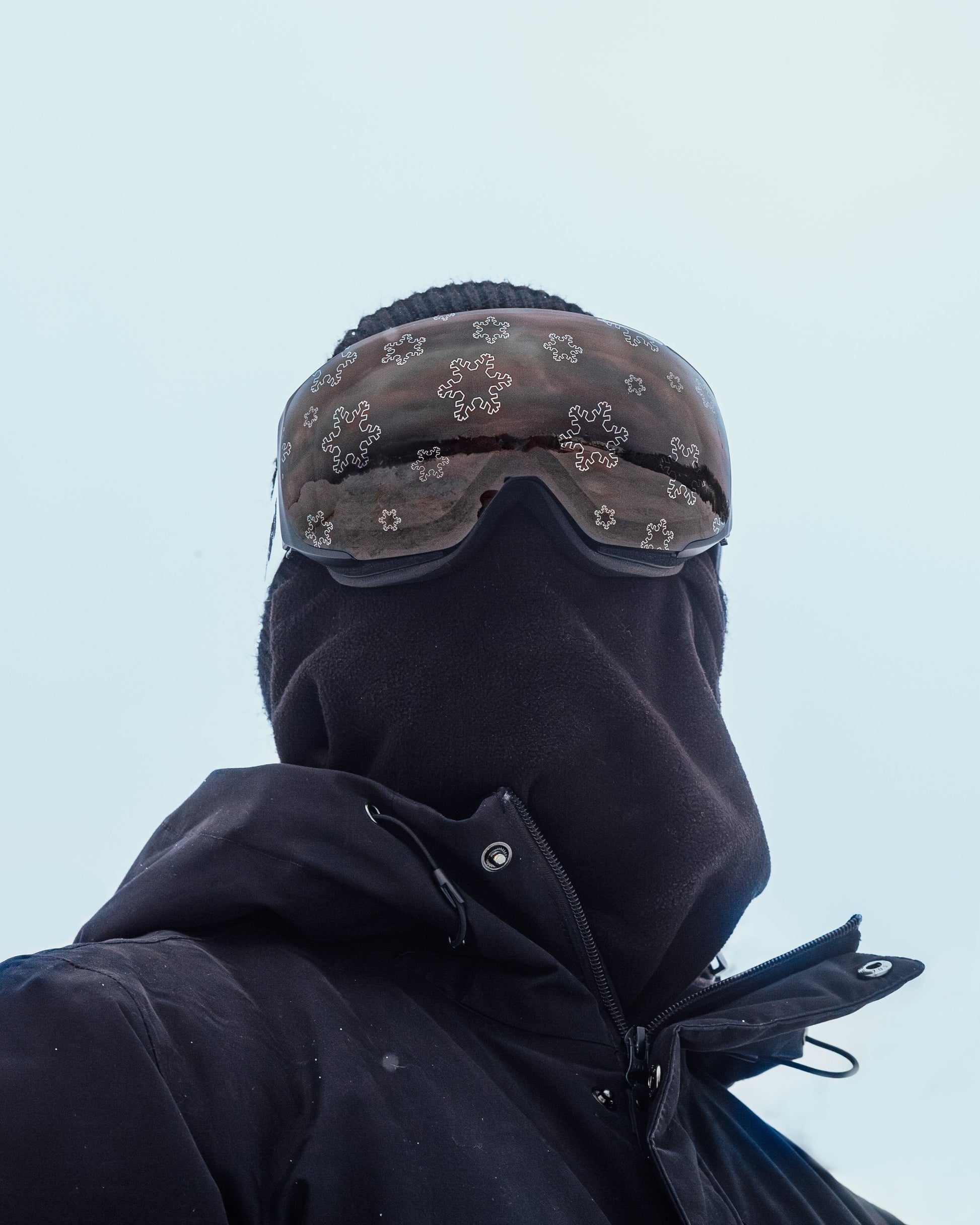 Skier wearing Skoggl's Winter Wonderland Lens ski goggles with silver snowflake designs on a dark lens, paired with an all-black winter outfit and full face covering. The minimalist, stealthy look highlights the high-performance, all-weather functionality of the goggles while showcasing bold snowflake details. Perfect for extreme conditions and alpine style.

