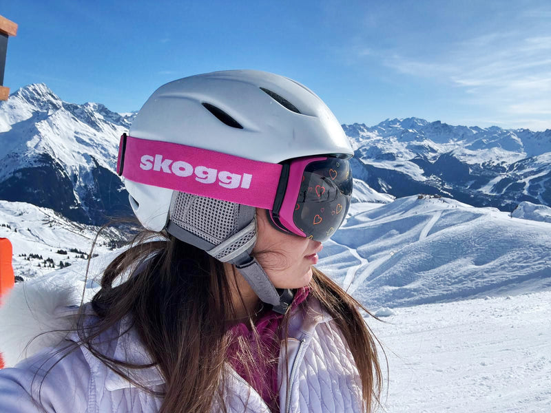A woman wearing pink Skoggl ski goggles with heart-shaped designs on the lens stands at the top of the Swiss Alps, showcasing the pink strap on the side as a snowy mountain range stretches behind her.