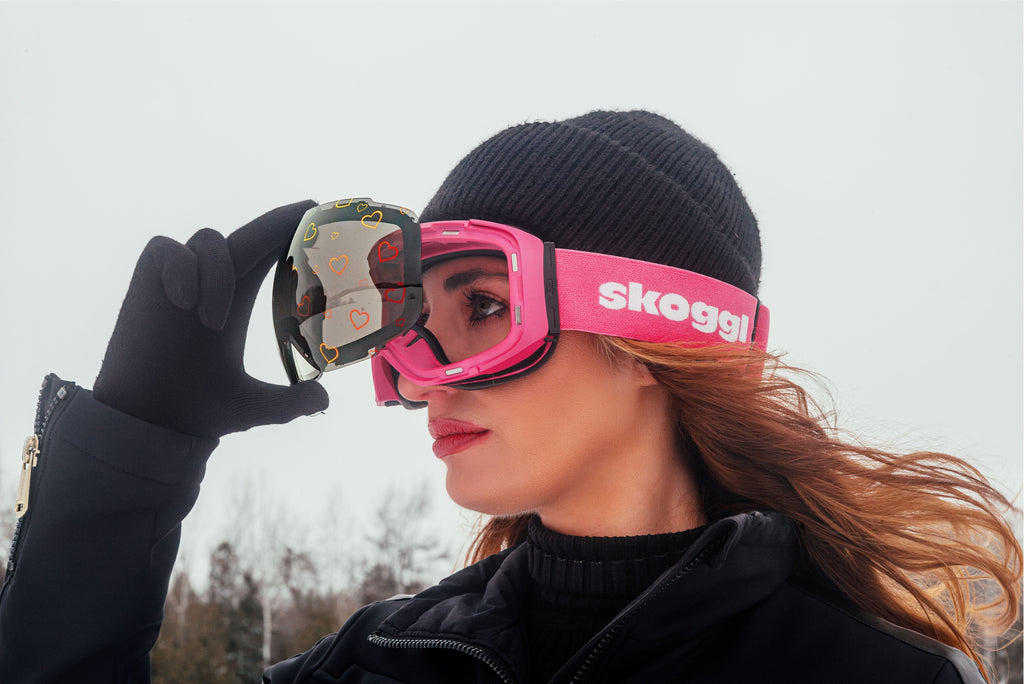 Female skier wearing pink Skoggl ski goggles with interchangeable magnetic heart-pattern lens on snowy mountain