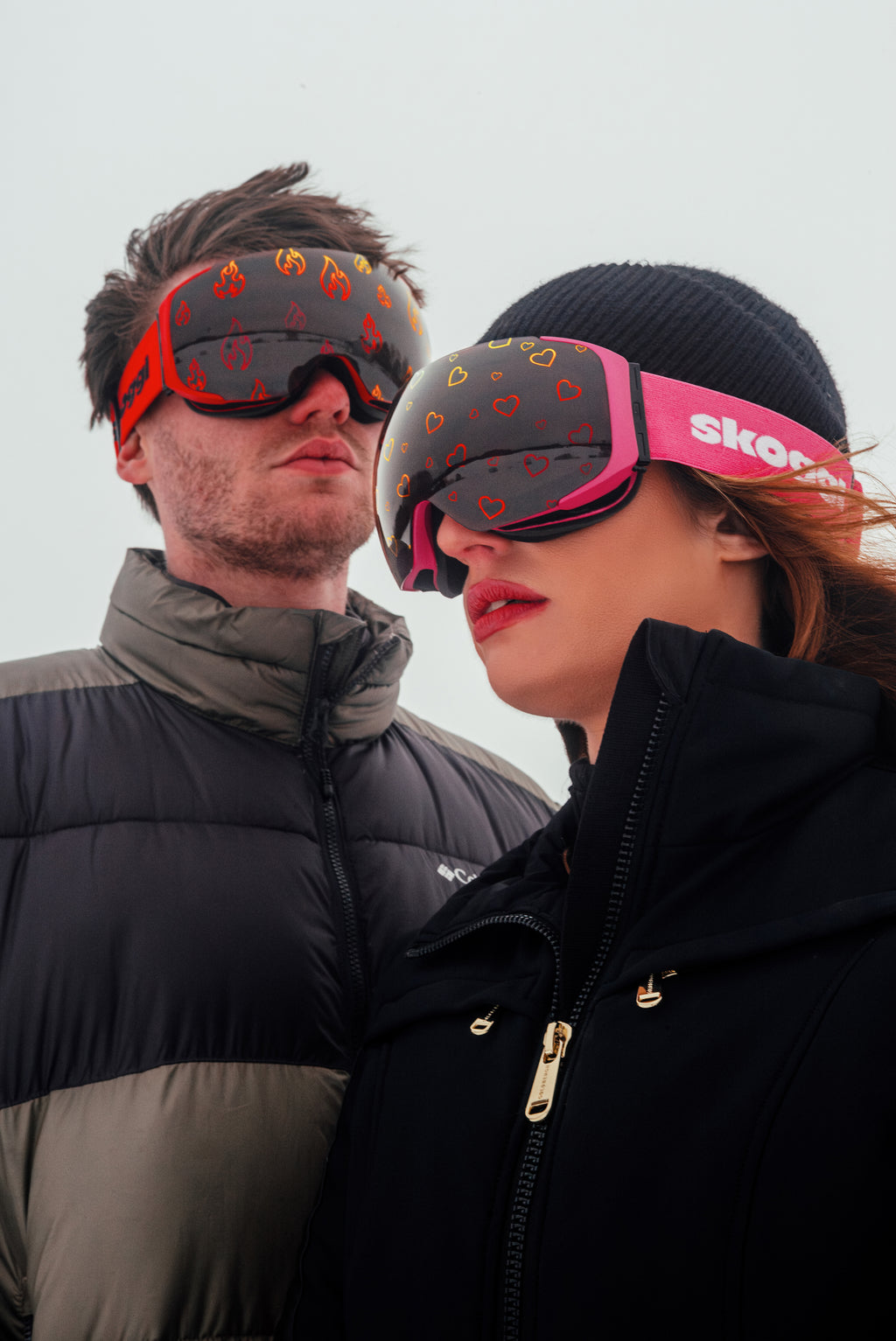Man and woman wearing Skoggl ski goggles with interchangeable magnetic lenses featuring flame pattern and heart pattern on snowy mountain