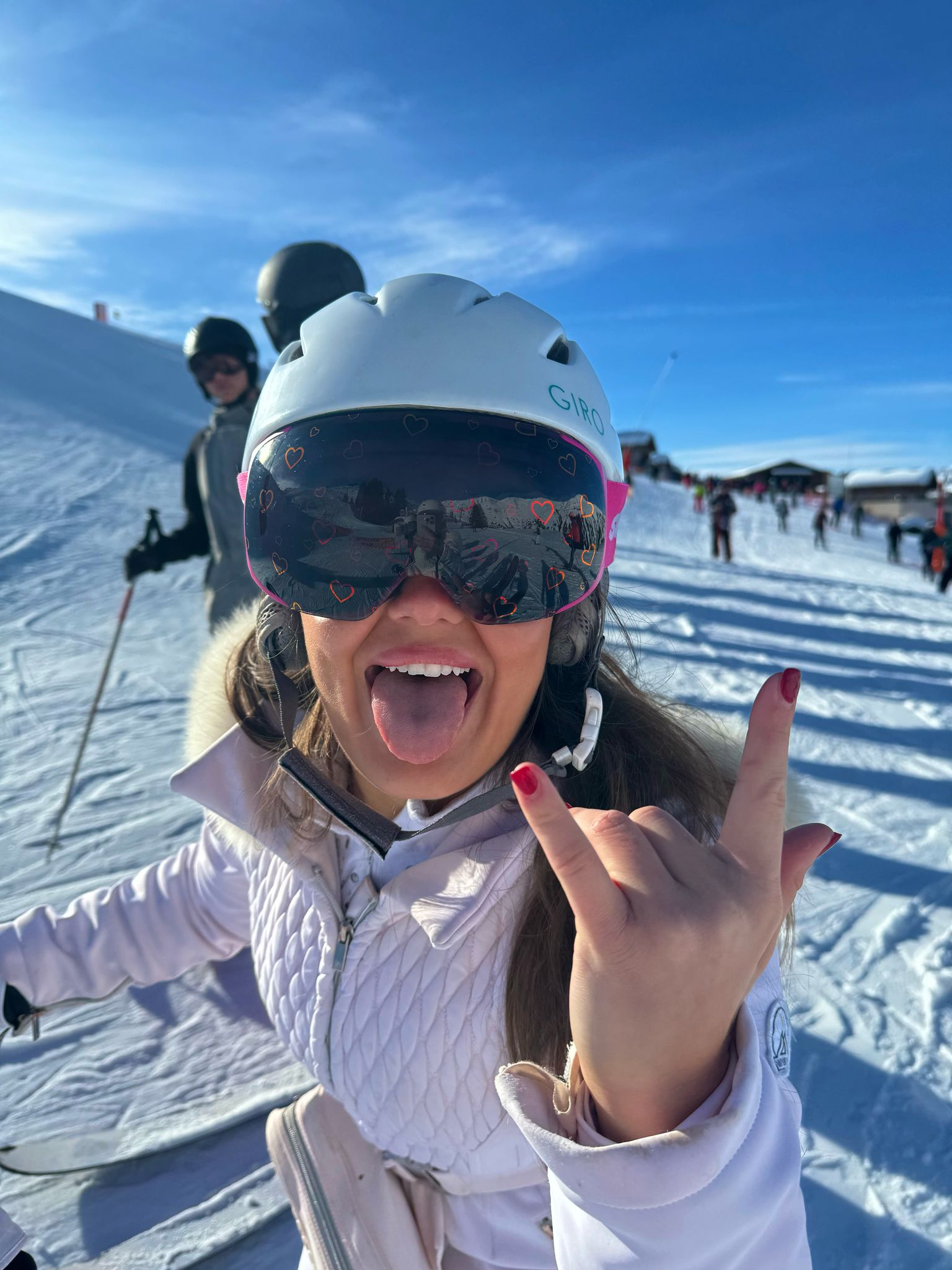 Person posing on a snowy slope wearing a helmet and pink Skoggl goggles with heart designs.