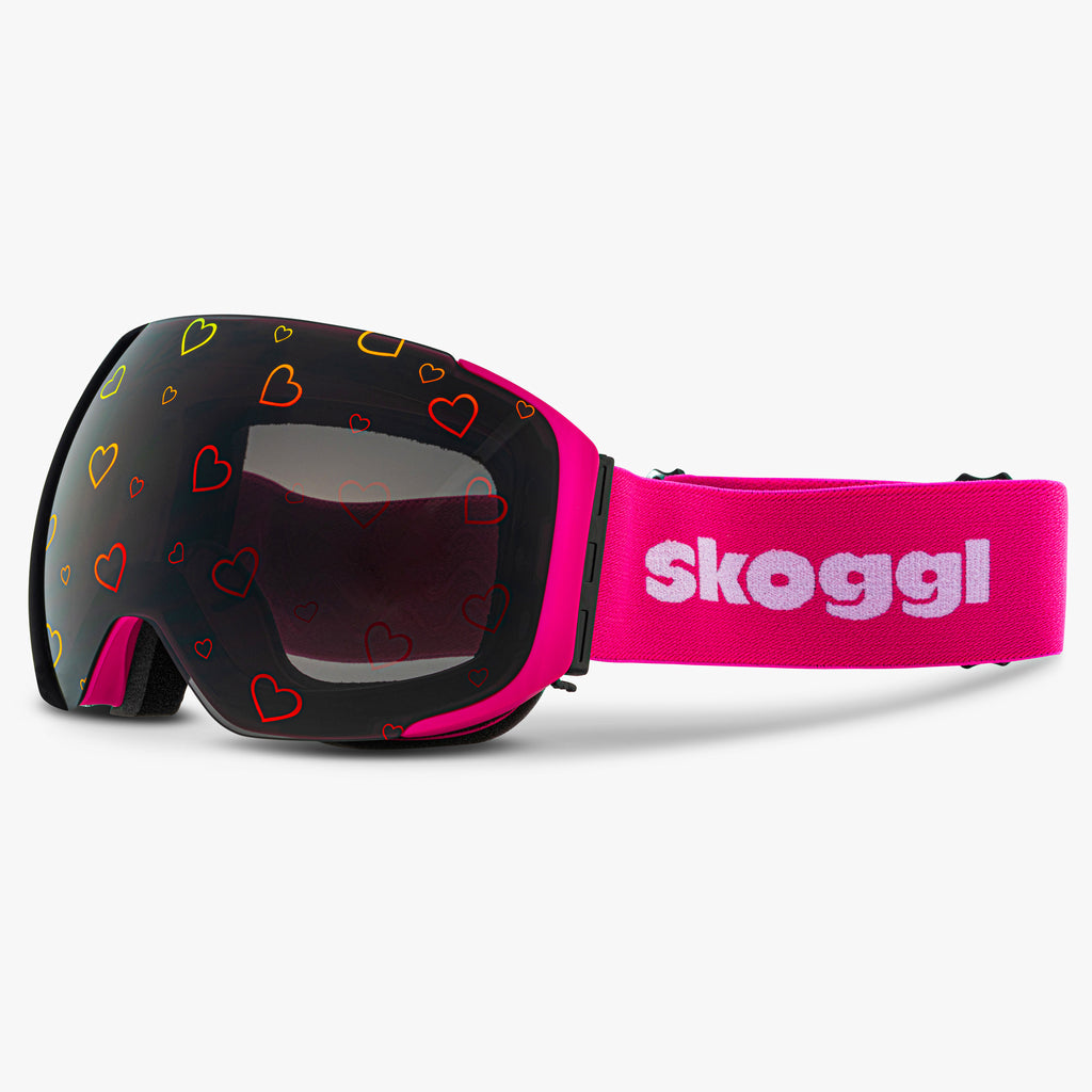 Diagonal view of Skoggl Lovefrost ski and snowboard goggles with hearts on the lens, featuring a pink frame, pink strap, and magnetic lens system for easy swaps. The playful heart design combines style and performance, making these goggles perfect for skiing, snowboarding, and winter sports.
