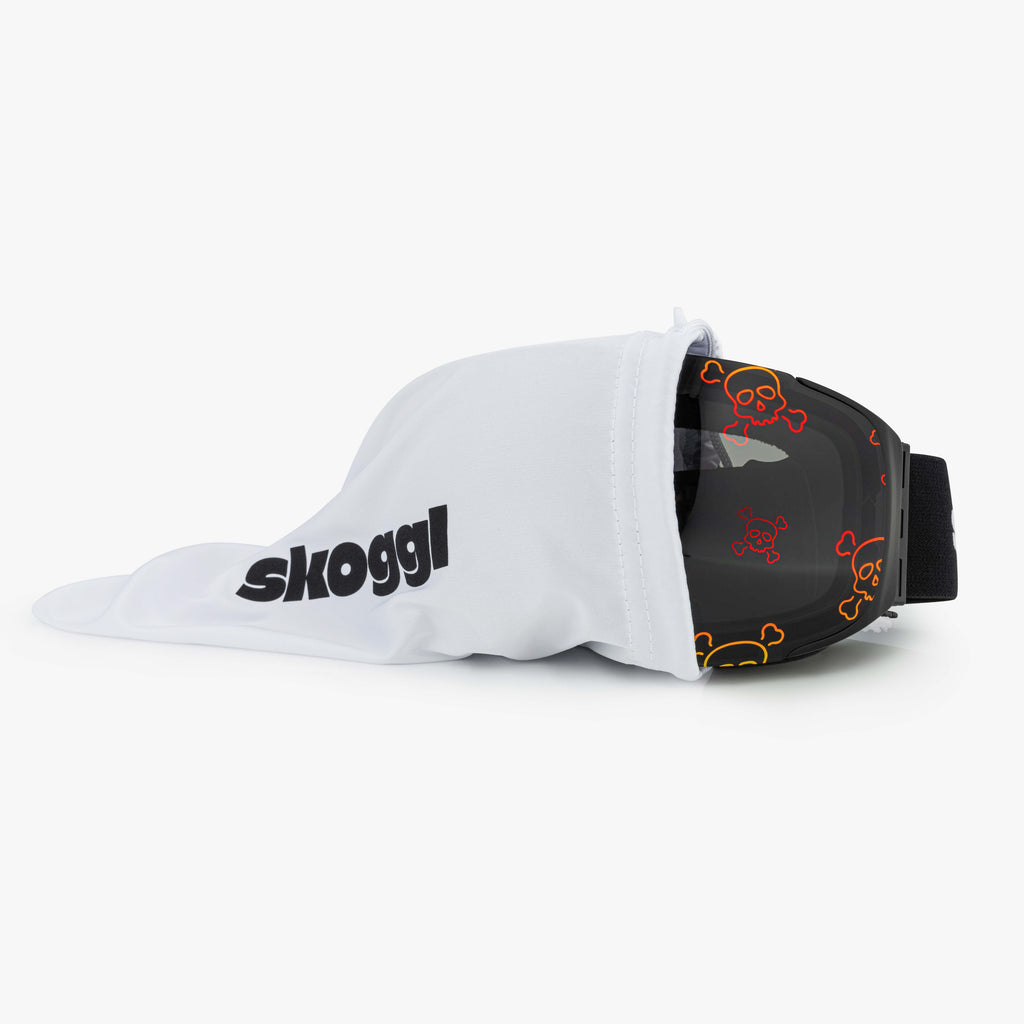 A pair of ski and snowboard goggles partially covered by a white dustbag with "Skoggl" written on it. The goggles feature a black strap and a lens with a bold red and yellow skull and crossbones pattern, showcasing edgy style and high-performance design for the slopes.