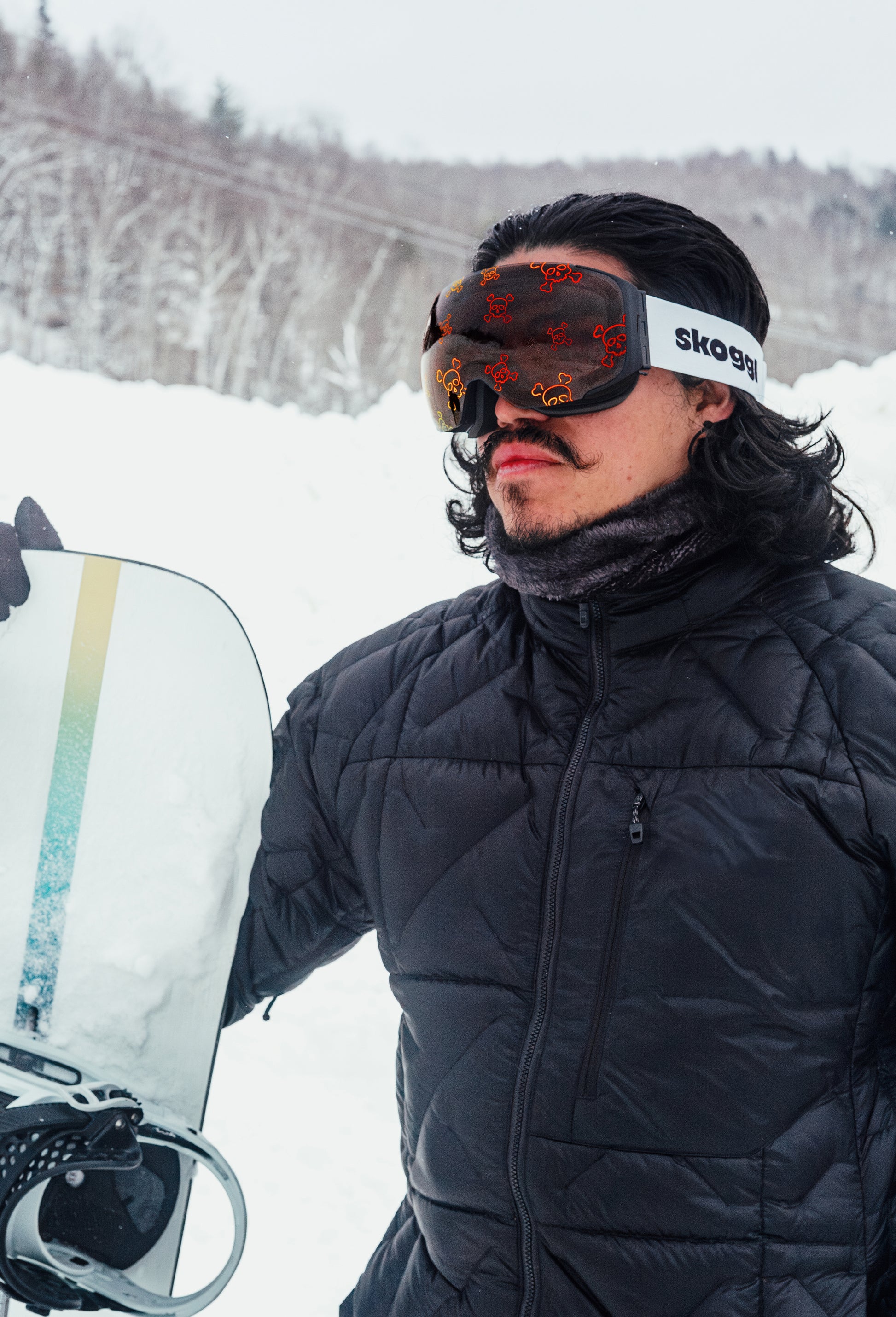 Model standing in the alps, posing with a snowboard and wearing Skoggl's Skully lens ski goggles featuring red skull and crossbones design.