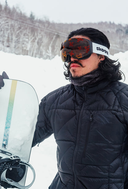 Model standing in the alps, posing with a snowboard and wearing Skoggl's Skully lens ski goggles featuring red skull and crossbones design.
