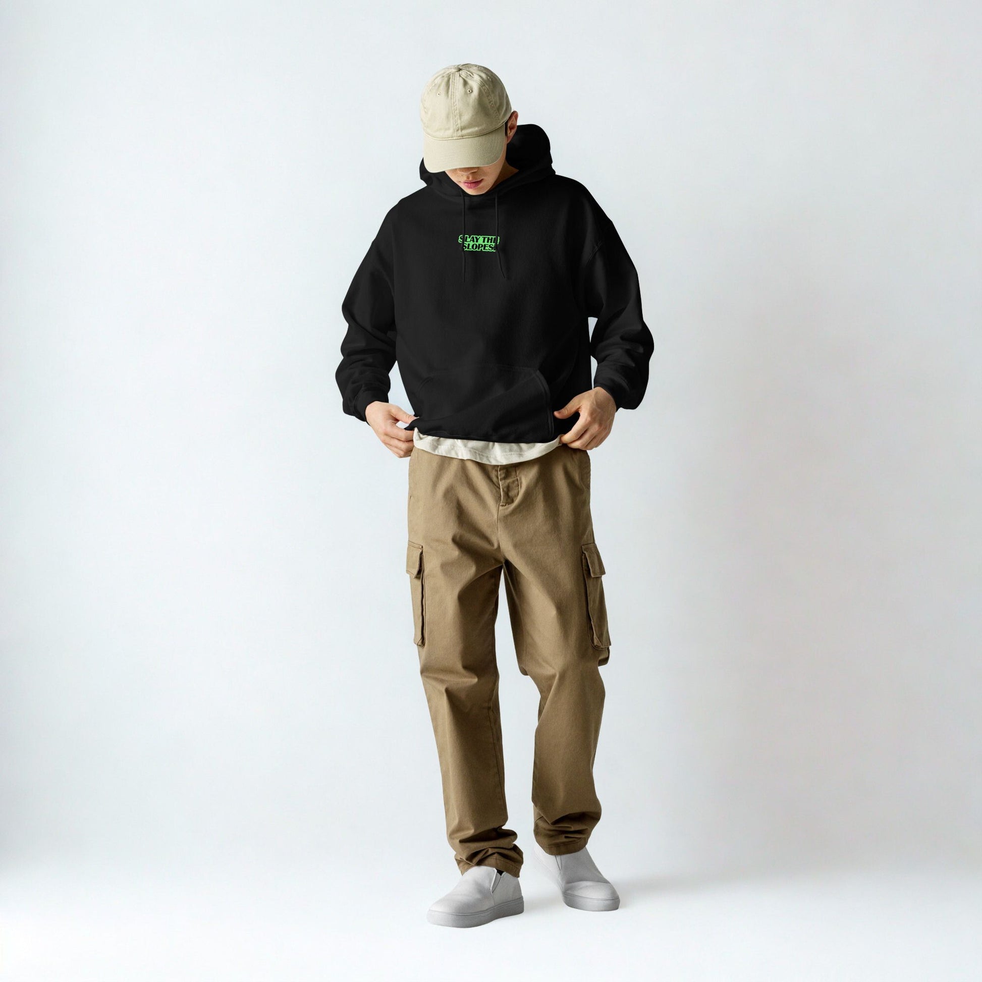 “Person dressed in a black Skoggl hoodie with bright green ‘Slay The Slopes’ text on the front, beige cap, and beige cargo pants, looking down. Streetwear-inspired winter outfit.