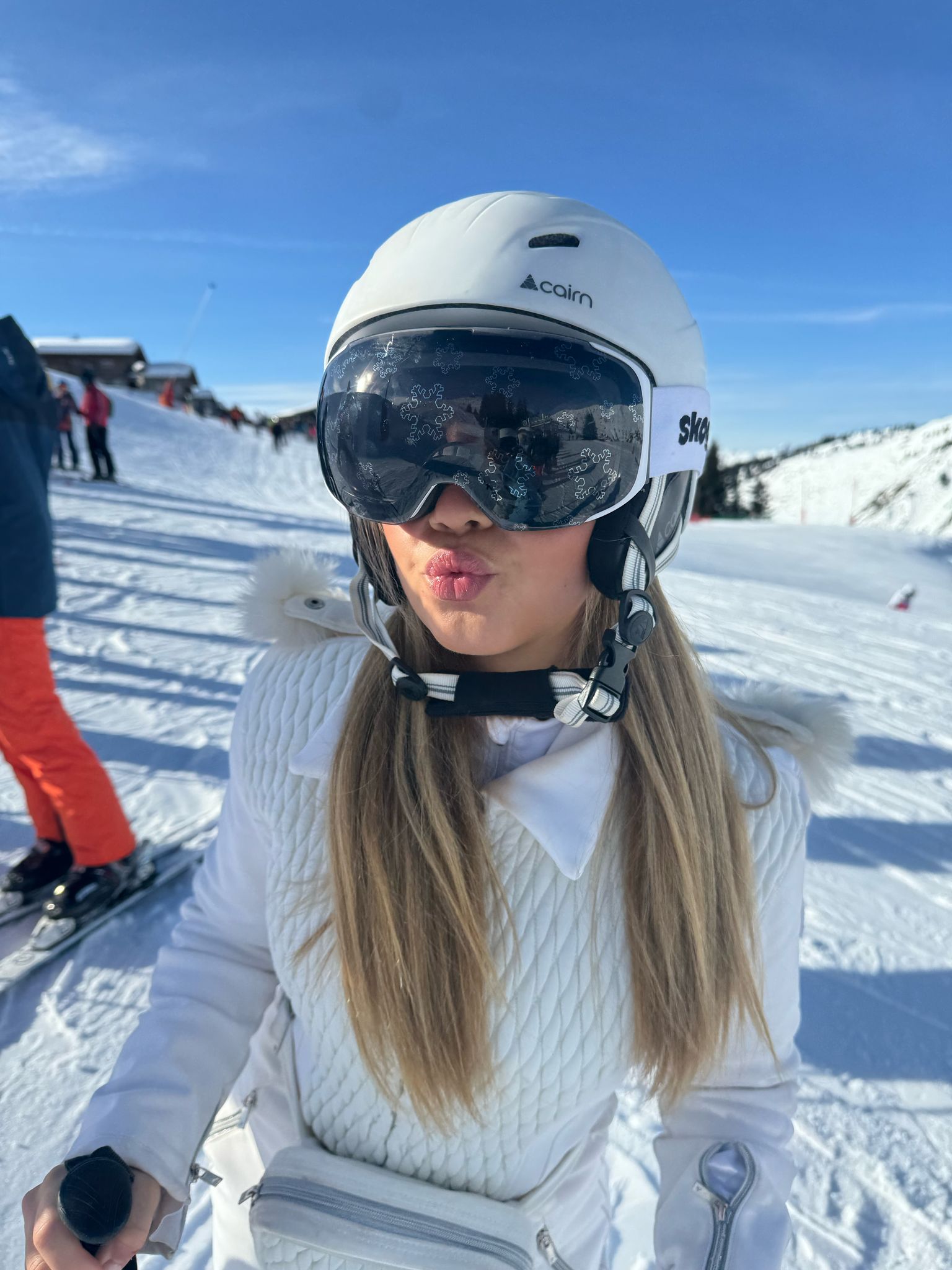 Person posing on a snowy slope wearing a helmet and white Skoggl goggles with snowflake designs.