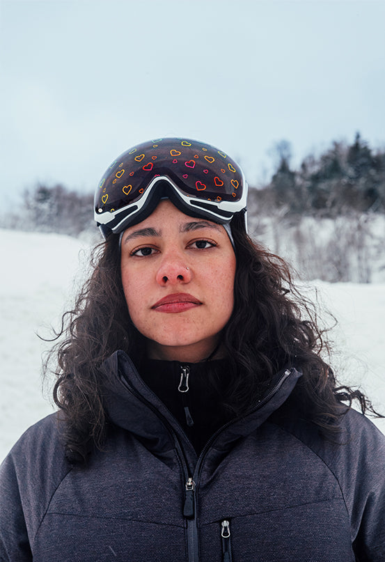 Close-up of a skier wearing Skoggl’s Sweetheart ski goggles, featuring a white frame and all-weather lens printed with heart designs. Shot outdoors against a snowy mountain backdrop, the model is dressed in a dark technical ski jacket, embodying a confident and stylish winter sports look. Ideal for showcasing fashionable, high-performance ski eyewear for women