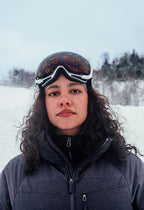 Close-up of a skier wearing Skoggl’s Sweetheart ski goggles, featuring a white frame and all-weather lens printed with heart designs. Shot outdoors against a snowy mountain backdrop, the model is dressed in a dark technical ski jacket, embodying a confident and stylish winter sports look. Ideal for showcasing fashionable, high-performance ski eyewear for women