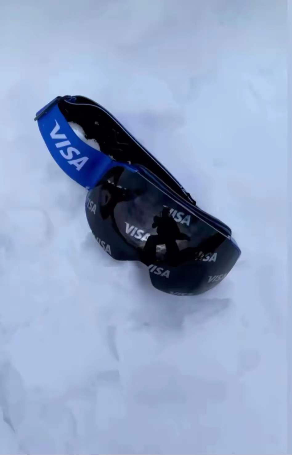 A pair of custom Skoggl  ski and snowboard goggles resting in soft white snow, created for a partnership with Visa. The goggles feature a bold royal blue strap with the white "VISA" logo and a dark mirrored lens that has the Visa logo subtly repeating across the surface.