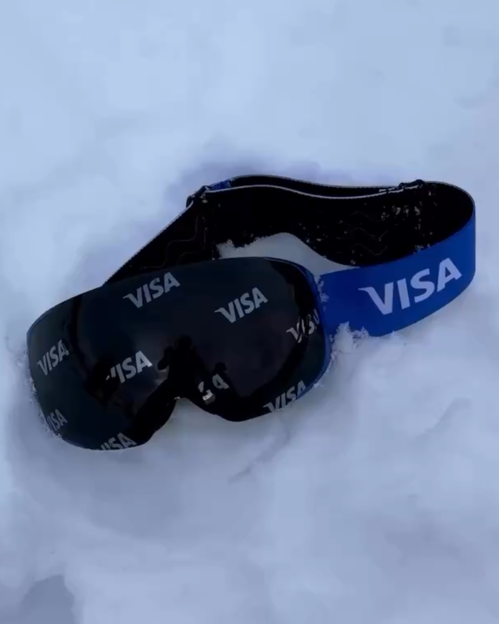 A pair of custom Skoggl  ski and snowboard goggles resting in soft white snow, created for a partnership with Visa. The goggles feature a bold royal blue strap with the white "VISA" logo and a dark mirrored lens that has the Visa logo subtly repeating across the surface.