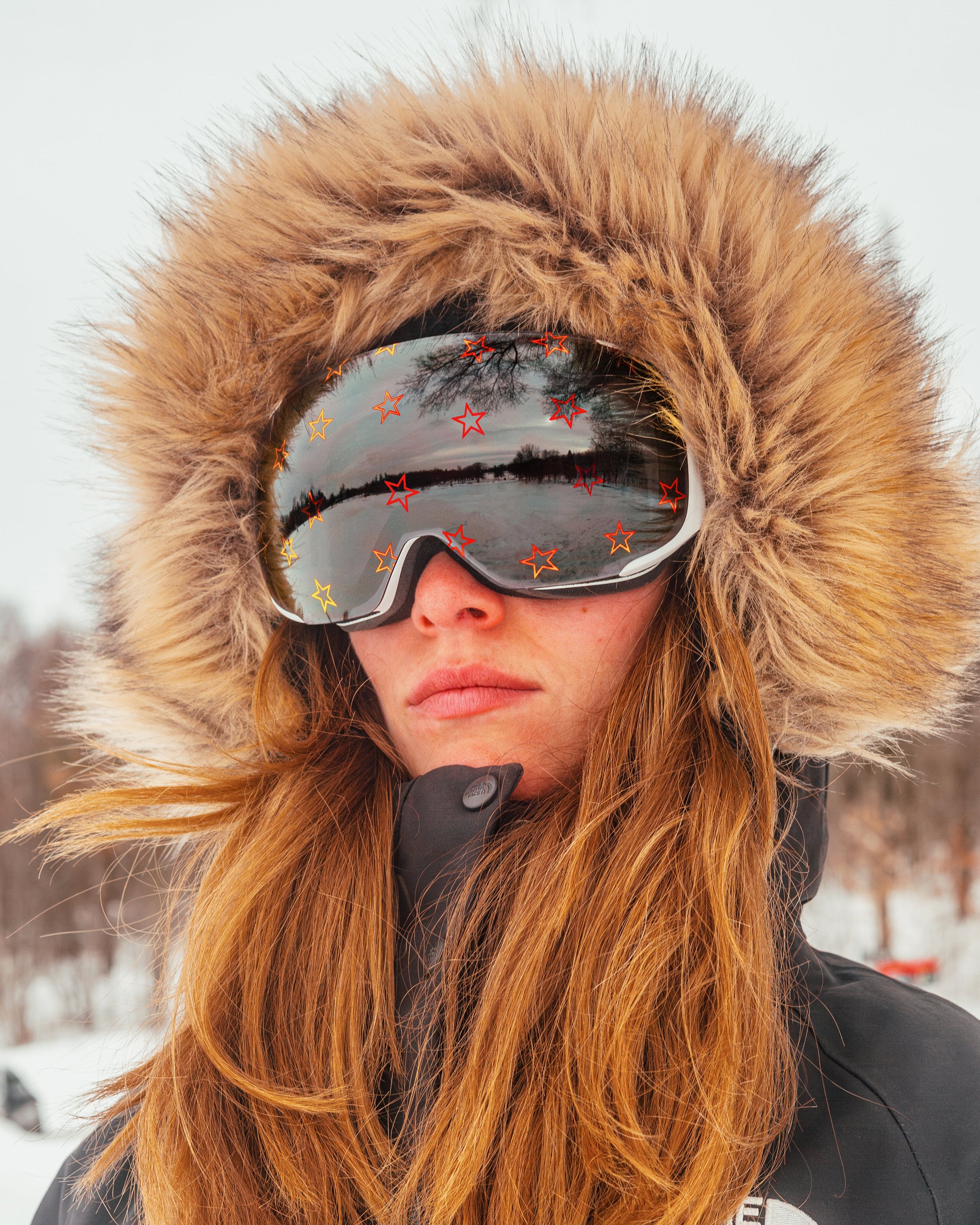 Female skier wearing Skoggl’s Star Gazer ski goggles with a white frame and all-weather lens printed with bold red and orange star designs. Styled in a fur-trimmed hood and black North Face jacket, she stands against a snowy landscape, showcasing premium performance eyewear designed for visibility and mountain style.