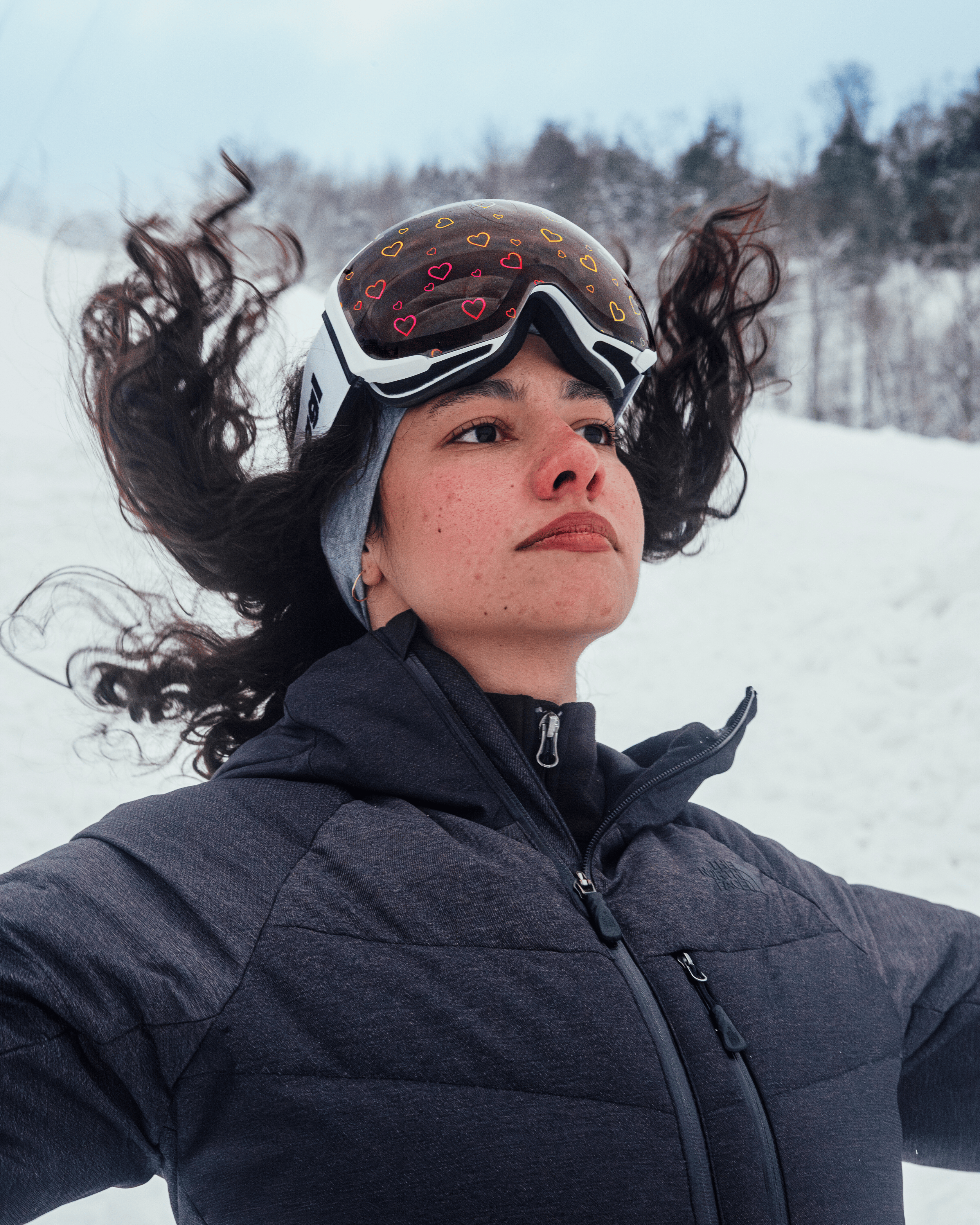 Woman wearing Skoggl ski goggles with heart-printed magnetic lens, standing confidently on snowy mountain – anti-fog, UV400 protection, stylish winter sports gear