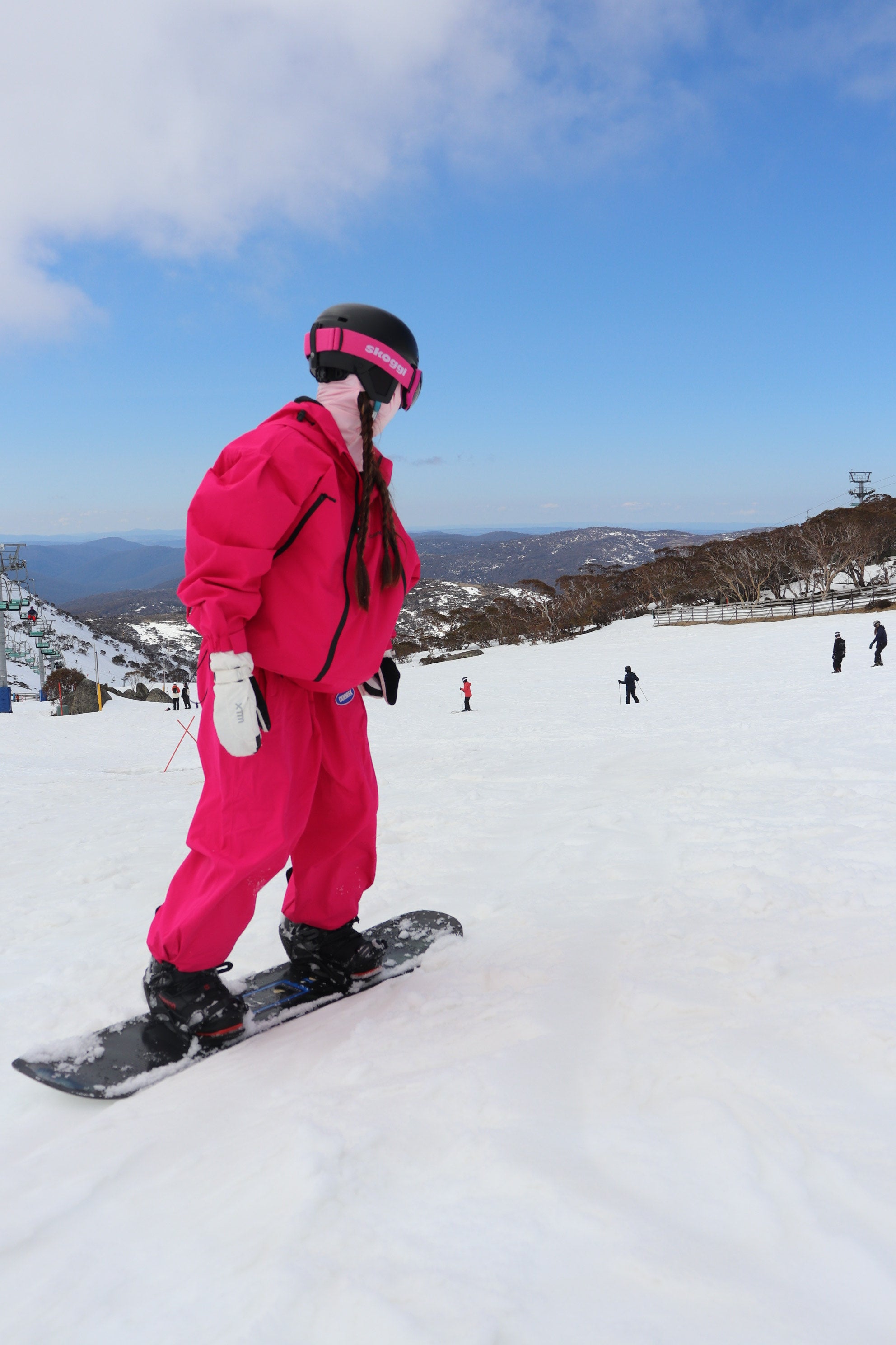 Person in pink snow gear wearing a helmet and Skoggl heart-design goggles, standing on a snowboard in a snowy landscape with mountains and a bright blue sky in australia.